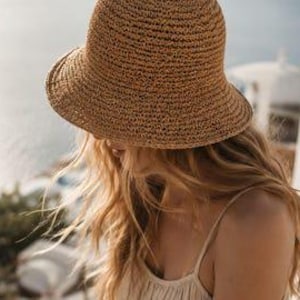 May include: A tan woven straw bucket hat with a rounded crown and a downward-sloping brim. The hat features a textured, open-weave construction made from natural fibres, providing a lightweight and flexible structure for sun protection.