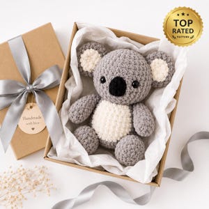 May include: A gray and white crocheted koala plush toy nestled inside a brown cardboard gift box lined with white tissue paper. The box features a gray ribbon bow and a round tag with the text Handmade with love and a heart symbol.