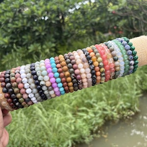 Può includere: Una collezione di numerosi braccialetti elastici con perline rotonde in pietre preziose levigate in un'ampia varietà di colori, tra cui sfumature di blu, rosa, verde, marrone, nero, bianco e arancione, esposti su un supporto cilindrico in juta beige.