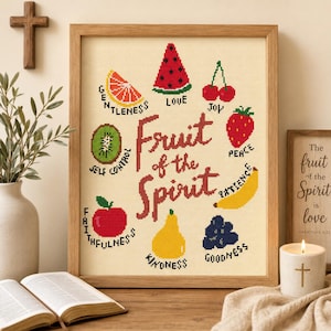 Op de afbeelding: Een ingelijst kruissteek kunstwerk op crèmekleurige stof met de tekst Fruit of the Spirit in donkerrood. Rondom de tekst staan kleurrijke geborduurde iconen van een grapefruit, watermeloen, kersen, aardbei, banaan, druiven, peer, kiwi en appel.