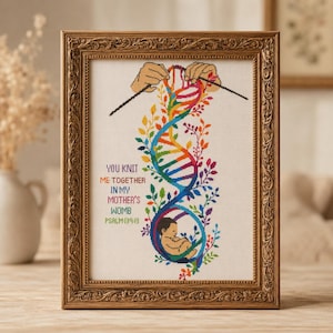 Può includere: Un ricamo a punto croce incorniciato che raffigura un filamento di DNA color arcobaleno lavorato a maglia da mani, con un feto alla base. Il testo recita: YOU KNIT ME TOGETHER IN MY MOTHER'S WOMB PSALM 139:13. La cornice è dorata, decorata e rettangolare.