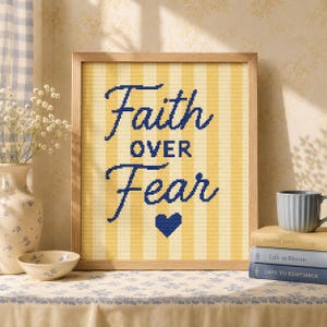May include: A framed cross-stitch embroidery featuring vertical yellow and cream stripes with the dark blue text Faith OVER Fear and a small dark blue heart icon centered below the lettering, all encased in a light-colored wooden frame.