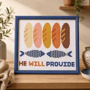 Op de afbeelding: Een ingelijst kruissteek kunstwerk met vijf verticale broden in de kleuren crème, roze, geel, oranje en bruin, geplaatst boven twee blauwe vissen. De tekst HE WILL PROVIDE is onderaan geborduurd in bruin, oranje en blauw.