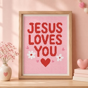 Op de afbeelding: Een ingelijst kruissteek borduurwerk met de rode tekst JESUS LOVES YOU op een lichtroze achtergrond. Het ontwerp bevat twee witte bloemen en verschillende kleine rode hartjes, gevat in een eenvoudige lichtgekleurde houten lijst.