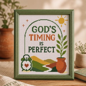 Op de afbeelding: Een ingelijst kruissteek borduurwerk met de tekst "GOD'S TIMING IS PERFECT" in donkergroen en oranje. Het ontwerp bevat een gele zon, een groene wekker met een rood hart, een plant in een pot en glooiende heuvels in groen en geel.