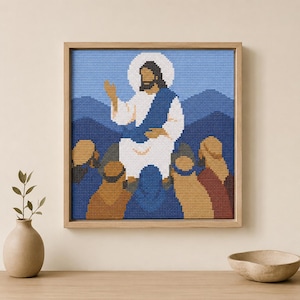 May include: A square cross-stitch embroidery in a light wood frame depicting a central figure in white robes with a blue sash and a white halo, surrounded by a group of people in tan, blue, and red robes against a backdrop of blue mountains.