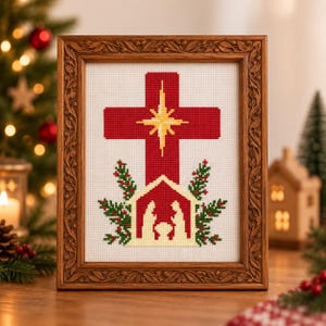 Könnte beinhalten: Ein gerahmtes Kreuzstichbild mit einem roten Kreuz und einem gelben Stern in der Mitte, platziert über einer roten Krippenszene. Das Motiv ist von grünen Tannenzweigen umgeben und befindet sich in einem dekorativen braunen Holzrahmen.