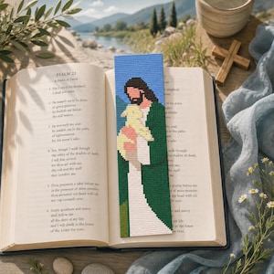 May include: A rectangular cross-stitch bookmark featuring a pixelated design of a figure with dark hair and a green robe holding a small yellow lamb. The bookmark is placed vertically on an open book displaying the text of Psalm 23 in English.