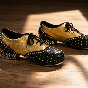 May include: A pair of tap shoes featuring a metallic gold upper and a black toe and heel section. The black portions are adorned with small gold studs. The shoes have black laces, a low heel, and a silver-toned metal tap plate on the sole.