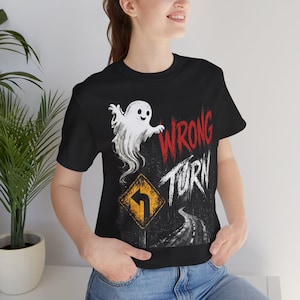 May include: A black short-sleeve t-shirt featuring a graphic print of a white ghost, a yellow diamond-shaped road sign with a black left-turn arrow, and the red and white text WRONG TURN above a dark, winding road illustration.