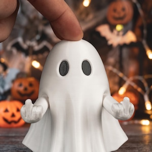 Può includere: Una statuetta di fantasma bianca con una testa liscia e arrotondata e due occhi ovali neri. Il fantasma ha due braccia articolate estese verso l'esterno, con ogni mano posizionata in modo da mostrare il gesto del dito medio.