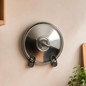 Puede incluir: Una tapa de olla circular de acero inoxidable está montada en la pared mediante dos soportes de plástico negro. La tapa presenta un asa central de metal y una superficie pulida y reflectante. Dos soportes sostienen la tapa por la parte inferior.