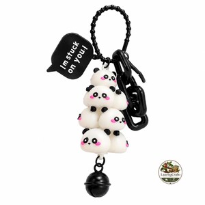 May include: A keychain featuring a stack of five white panda figures with black ears, eyes, and pink cheeks. A black speech bubble charm with the white text 'I'm stuck on you!' is attached to a black ring, along with a black bell at the bottom.