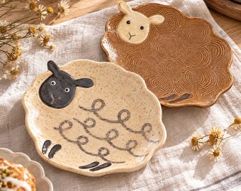 May include: Two ceramic plates shaped like sheep. One plate is cream-colored with a black sheep head and dark gray curly line details. The other plate is brown with a cream-colored sheep head and embossed spiral patterns across the surface.