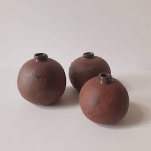 Op de afbeelding: Drie bolvormige keramische vazen met een matte, donker roodbruine afwerking en een textuur op het oppervlak. Elke vaas heeft een kleine, smalle, donkerbruine cilindrische hals aan de bovenkant. De vazen staan in een groep op een witte ondergrond.