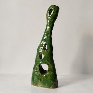 Op de afbeelding: Een hoge, met de hand gevormde keramische sculptuur met een glanzend, diepgroen glazuur. Het object heeft een onregelmatige, langwerpige vorm met vier opvallende, afgeronde openingen van verschillende groottes die verticaal zijn geplaatst.