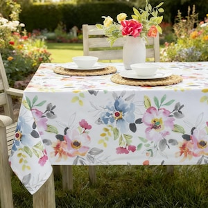 May include: A white rectangular tablecloth featuring a watercolor floral pattern in shades of pink, blue, yellow, and grey. The table is set with two white bowls on woven placemats and a white vase containing a bouquet of pink and yellow flowers.