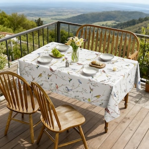 May include: A white rectangular tablecloth featuring a colorful pattern of various birds perched on thin brown branches with green leaves. The tablecloth is draped over a wooden table set with white plates, gold cutlery, and a glass vase of flowers.