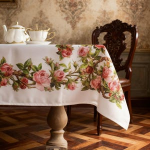 May include: A white tablecloth featuring a horizontal border print of pink roses, green leaves, and thorny stems. The fabric drapes over a table, displaying a detailed floral pattern with various stages of rose blooms across the white surface.