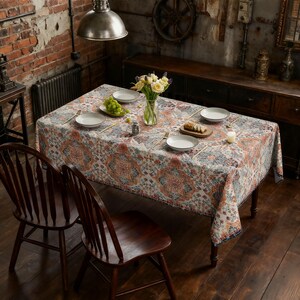 May include: A rectangular tablecloth featuring a repeating intricate floral and geometric pattern in shades of orange, blue, and cream. The cloth covers a dining table set with white plates, a glass vase of yellow flowers, and a small wooden board.