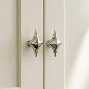 Puede incluir: Dos pomos de armario en tono plateado pulido con una forma distintiva de estrella de cuatro puntas o diamante. Estas piezas de herraje metálico están montadas en dos puertas de armario de color blanco roto, con una superficie lisa y brillante.
