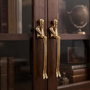 May include: Two polished gold-toned metal cabinet handles shaped like stylized figures sitting with their legs extending downward. The figures have smooth, reflective surfaces and are mounted vertically on the glass doors of a dark wood cabinet.