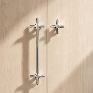 Op de afbeelding: Een lange, verticale kastgreep van gepolijst chroom en een bijpassende, kleinere kastknop, beide in de vorm van een vierpuntige ster met afgeronde randen, gemonteerd op lichtgekleurde houten kastdeuren met een zichtbare houtnerf.