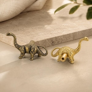 May include: Two dinosaur-shaped cabinet knobs with a metallic gold finish. Each knob features a long-necked sauropod design with textured skin details. One knob is shown from the side, while the other displays the cylindrical mounting post.