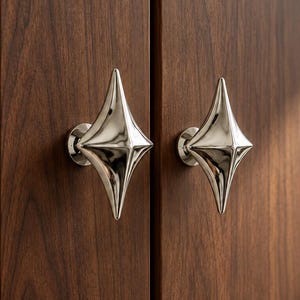 Puede incluir: Dos pomos de armario de metal plateado pulido con forma de estrella de cuatro puntas fijados en puertas de armario de madera de color marrón oscuro. La palabra XUTIA está impresa en la esquina inferior derecha de la imagen en una fuente blanca simple.