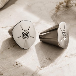 May include: A pair of silver-toned metal earrings with a polished finish. Each earring features a flat, irregular circular face engraved with a central spiral design surrounded by four radiating straight lines, resembling a stylized sun motif.