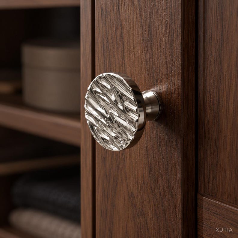 May include: A circular cabinet knob with a polished silver finish and a textured, rippled surface. The knob is mounted on a dark brown wooden cabinet door. The word XUTIA is printed in small white letters in the bottom right corner.