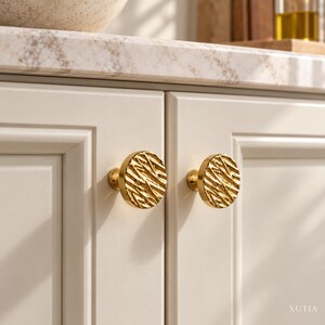May include: Two round, gold-toned cabinet knobs with a textured, wavy, and ridged surface pattern. The knobs are mounted on light-colored, paneled cabinet doors. The text XUTIA is visible in the bottom right corner of the image.