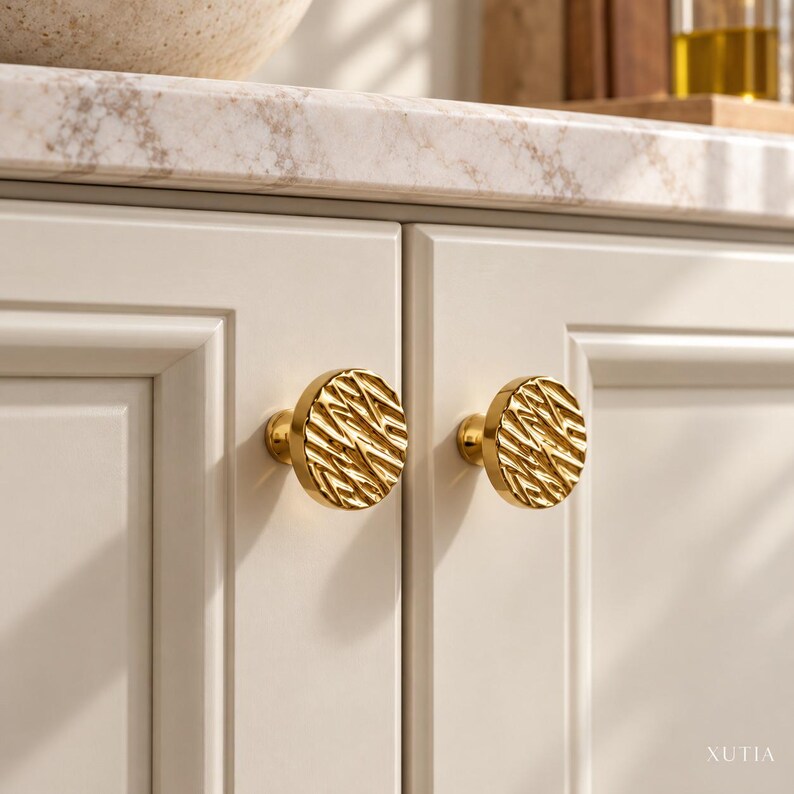 May include: Two round, gold-toned cabinet knobs with a textured, wavy, and ridged surface pattern. The knobs are mounted on light-colored, paneled cabinet doors. The text XUTIA is visible in the bottom right corner of the image.