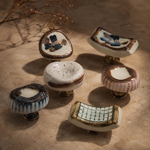 May include: A collection of six ceramic cabinet knobs with brass-toned bases. The knobs feature various shapes, including round and rectangular forms, decorated with hand-painted patterns such as blue flowers, stripes, speckles, and grid designs.