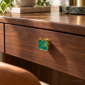 May include: A clover-shaped drawer knob featuring a vibrant green marbled resin inlay encased in a polished gold-tone metal frame, mounted on a dark brown wood drawer front with the text XUTIA visible in the bottom right corner.