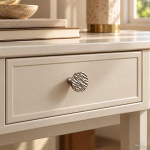 May include: A round, silver-toned cabinet knob with a textured, organic surface pattern attached to a light cream-colored wooden drawer. The knob features a series of irregular, raised lines and grooves that create a reflective, metallic finish.