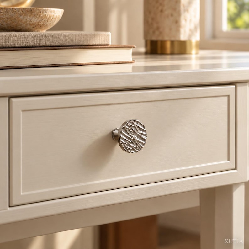 May include: A round, silver-toned cabinet knob with a textured, organic surface pattern attached to a light cream-colored wooden drawer. The knob features a series of irregular, raised lines and grooves that create a reflective, metallic finish.