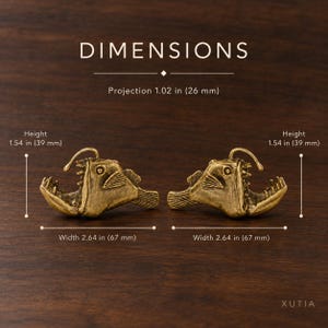 Op de afbeelding: Twee messingkleurige beslagstukken in de vorm van een zeeduivel. De tekst luidt DIMENSIONS, Projection 26 mm, Height 39 mm, Width 67 mm. Elk stuk toont een gedetailleerde vissenkop met scherpe tanden en een rugvin, afgebeeld vanaf de zijkant.