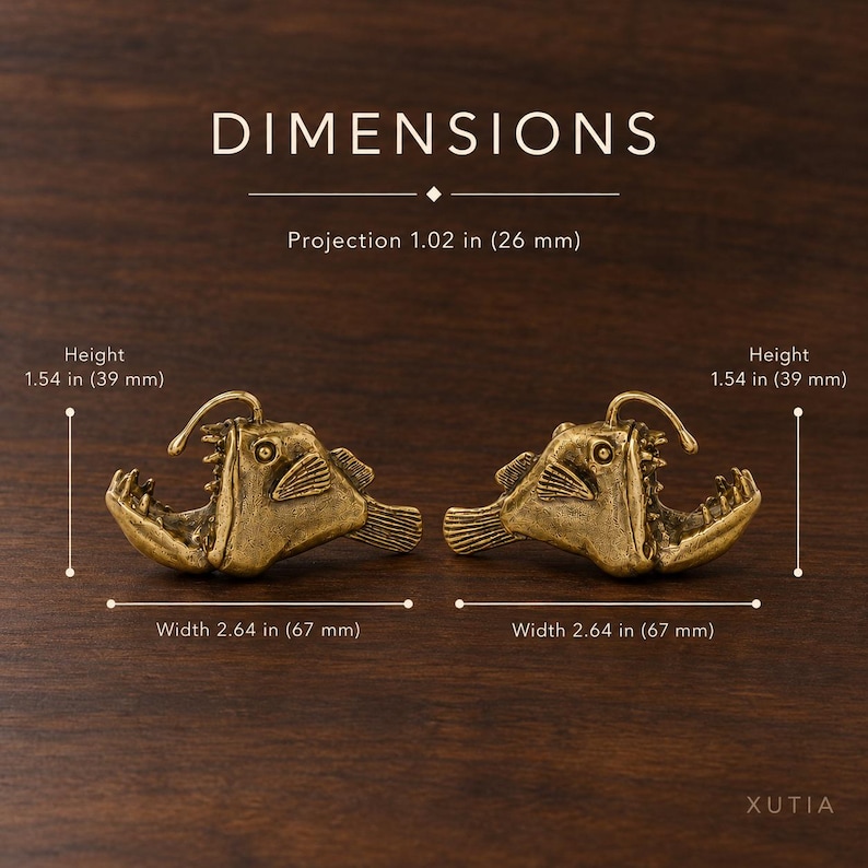 Op de afbeelding: Twee messingkleurige beslagstukken in de vorm van een zeeduivel. De tekst luidt DIMENSIONS, Projection 26 mm, Height 39 mm, Width 67 mm. Elk stuk toont een gedetailleerde vissenkop met scherpe tanden en een rugvin, afgebeeld vanaf de zijkant.