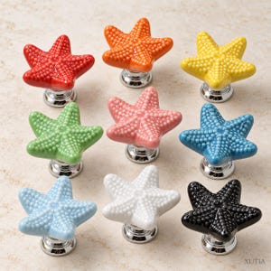 May include: A collection of nine ceramic drawer knobs shaped like starfish, each featuring a textured, bumpy surface and a polished silver-tone metal base. The knobs are available in red, orange, yellow, green, pink, blue, light blue, white, and black.