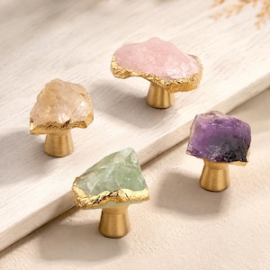 May include: Four cabinet knobs featuring raw, irregularly shaped gemstone tops in pale pink, light green, translucent white, and deep purple. Each stone is accented with a metallic gold-toned edge and mounted on a conical brass-colored base.