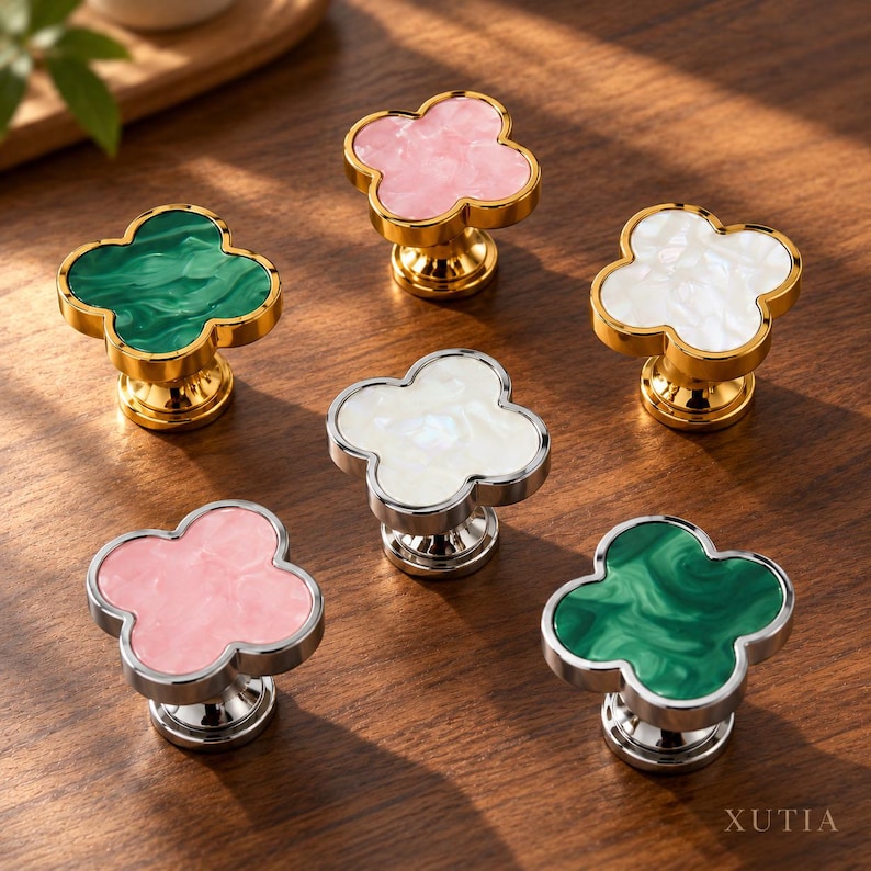 May include: Six clover-shaped cabinet knobs with metallic bases and marbled resin inserts in pink, green, and white. Three knobs feature gold-tone bases, while three feature silver-tone bases. The word XUTIA is printed in the bottom right corner.