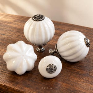 May include: Four white ceramic cabinet knobs with metallic hardware. Two knobs are spherical and ribbed with dark metal caps, one is a smooth round knob with a silver Celtic knot inlay, and one is a fluted, star-shaped knob. Text reads XUTIA.