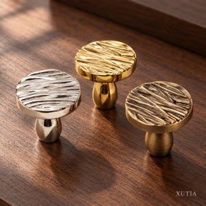 May include: Three cabinet knobs with a round, textured top and a smooth, tapered base. One knob is polished silver, one is polished gold, and one is a brushed bronze finish. The word XUTIA is printed in the bottom right corner.