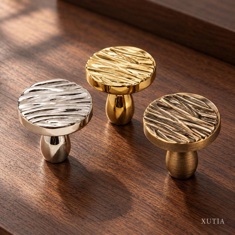 May include: Three cabinet knobs with a round, textured top and a smooth, tapered base. One knob is polished silver, one is polished gold, and one is a brushed bronze finish. The word XUTIA is printed in the bottom right corner.