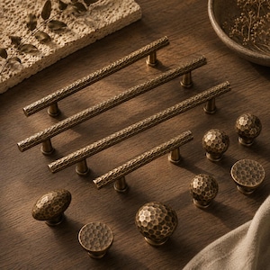 May include: A collection of antique brass cabinet hardware featuring four long, slender bar pulls and seven round knobs. Each piece displays a consistent hammered, textured finish across its surface, providing a tactile and uniform appearance.
