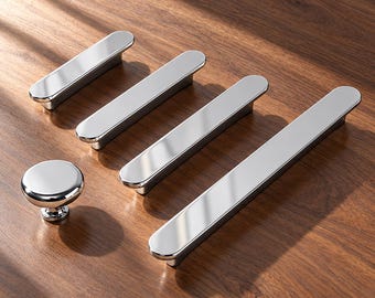 Könnte beinhalten: Ein Set aus polierten Chrom-Möbelbeschlägen, bestehend aus einem runden Knopf und drei länglichen, rechteckigen Griffen in verschiedenen Längen, alle mit einer glatten, reflektierenden metallischen Oberfläche.