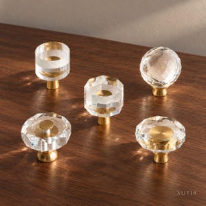 May include: Five cabinet knobs featuring clear acrylic tops and brushed brass bases. The set includes various geometric shapes: a cylindrical knob, a faceted sphere, and three flat-topped hexagonal designs with central brass accents. The text XUTIA is visible.