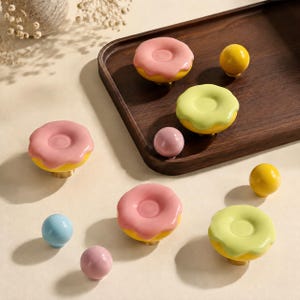 Op de afbeelding: Een collectie kastknoppen waaronder diverse in de vorm van donuts met roze of lichtgroen glazuur op gele basissen, naast kleinere bolvormige knoppen in effen gele, lichtblauwe en roze kleuren, gerangschikt op een houten dienblad.