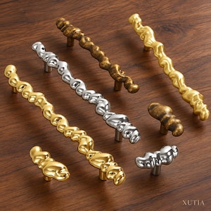 May include: A collection of cabinet hardware including long, irregular, organic-shaped pulls and matching knobs in polished gold, silver, and antique bronze finishes. The metal pieces feature a textured, molten-like surface. The text XUTIA is visible.